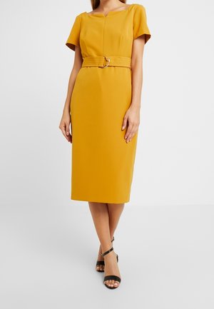 Femme portant une robe jaune moutarde mi-longue aux genoux avec manches courtes, taille ceinturée et talons ouverts noirs, debout sur un fond blanc.