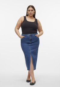 Top sin mangas negro y falda de mezclilla azul lápiz con abertura frontal. La falda cuenta con bolsillos y una cintura. La modelo está de pie con las manos en los bolsillos.