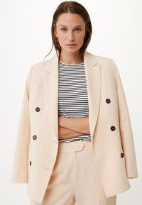 Blazer beige doppiopetto con grandi bottoni marroni, indossato sopra una camicia a righe bianche e nere. Tessuto liscio, vestibilità sagomata e rever definiti.