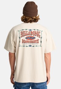 Beige cotton t-shirt with a large retro-style graphic reading "BILLABONG BOARDSHORTS" in red, accented by green waves and borders.