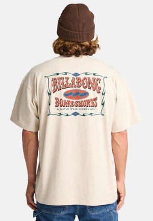 Beige katoenen t-shirt met een grote retro-stijl afbeelding met de tekst "BILLABONG BOARDSHORTS" in het rood, omlijst door groene golven en randen.