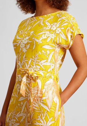Jumpsuit floral jaune avec une coupe ample, des manches courtes, une taille cintrée avec une ceinture nouée, et des motifs de grues en blanc et orange.