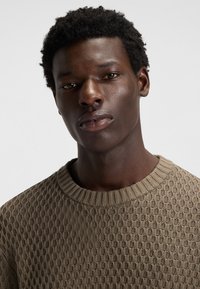 Junger schwarzer Mann mit kurzen lockigen Haaren, der einen strukturierten braunen Strickpullover trägt, frontal blickend mit einem neutralen Ausdruck vor einem schlichten Hintergrund.