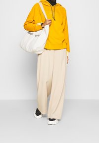 Gul hoodie med framficka, beige vida byxor och svarta träningsskor. Accessoar med en vit tygväska med text.