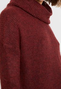 Sweter w głębokim odcieniu czerwieni, z dekoldem typu cowl, prążkowaną fakturą oraz luźnym fasonem. Materiał wydaje się miękki i ciepły.