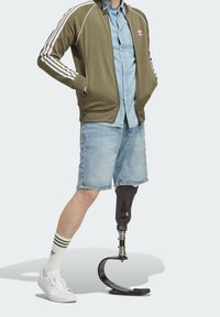 Mężczyzna w oliwkowo-zielonej kurtce zapinanej na zamek, dżinsowej koszuli i dżinsowych szortach, z czarną protezą nogi i białymi tenisówkami.