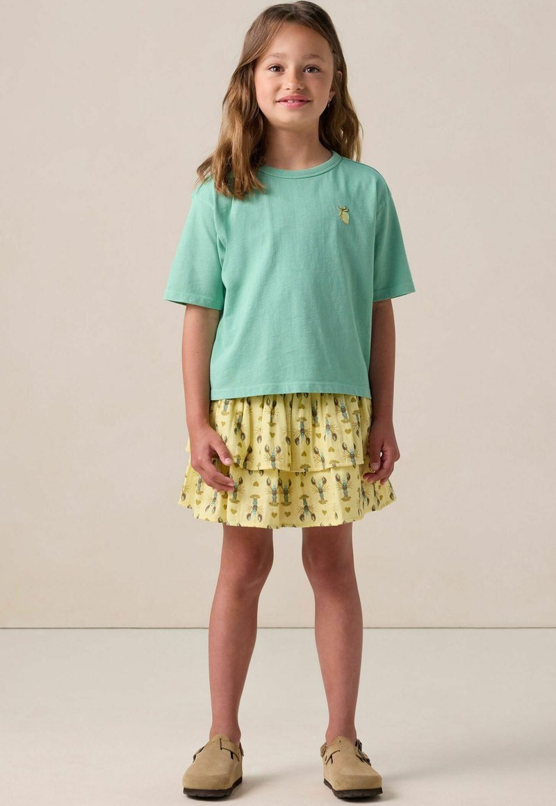 T-shirt vert menthe à manches courtes avec un petit motif jaune, associé à une jupe jaune à volants présentant un motif de homard et des chaussures marron.