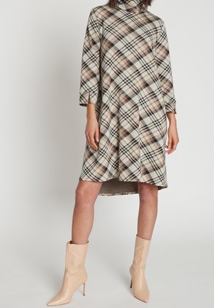Vrouw draagt een beige, bruine en zwarte geruite jurk met hoge hals en driekwart mouwen, en beige enkellaarzen met hoge hak.