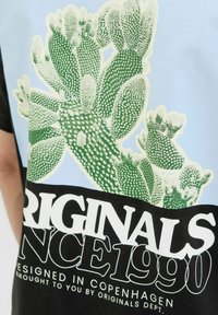 Must tüüpi must ja helesinine graafiline T-särk, millel on roheline kaktuse print ja allosas bold-kirjas "ORIGINALS SINCE 1990" tekst valgete tähtedega.