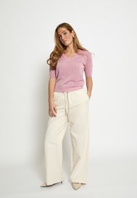 Maglione rosa a maniche corte con scollo a V in materiale scintillante abbinato a pantaloni color crema a vita alta e gamba larga, con tacchi beige appuntiti.