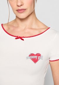 Hvit t-skjorte med korte ermer, rød blondekant og et hjerteformet grafisk motiv som har "tommy girl" i sølvfarge og rhinestones.