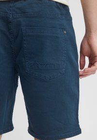Pantalones cortos azul marino en una tela ligera que presenta un bolsillo trasero con un diseño bordado y cierre de botón. Textura suave, corte entallado.