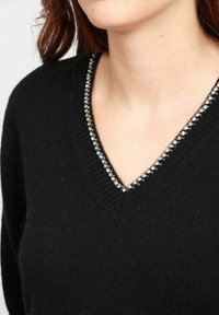 Donna che indossa un maglione nero a V con un bordo di strass argento lungo il colletto, con il collo e parte dei capelli visibili.