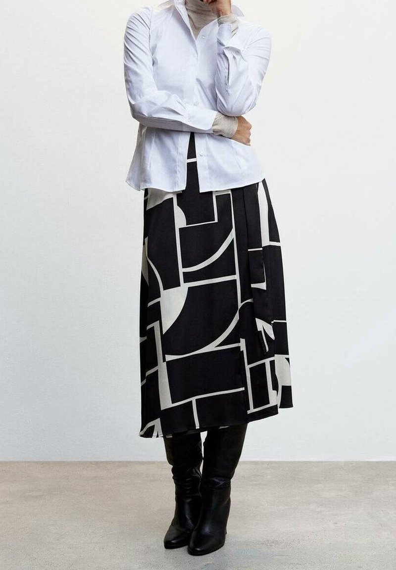 Personne portant une chemise blanche boutonnée, une jupe midi géométrique noire et blanche et des bottes noires montantes, debout sur un sol uni.