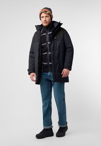 Schwarze Daunenjacke mit grauen Akzenten, Reißverschlusstaschen und einer blau gestreiften Strickmütze. Getragen mit einem gemusterten Fleece-Pullover und blauen Jeans.