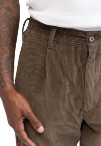 Pantalons en velours côtelé marron foncé avec des côtes verticales. Dotés d'une fermeture à bouton et de poches latérales. Le tissu a une texture douce.