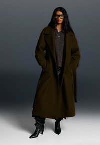 Tmavozelený dlhý kabát so strihom do rovna, širokými klopami, bočnými vreckami, položený na šedom textúrovanom svetri a čiernych kožených nohaviciach, doplnený o čižmy na vysokom opätku.