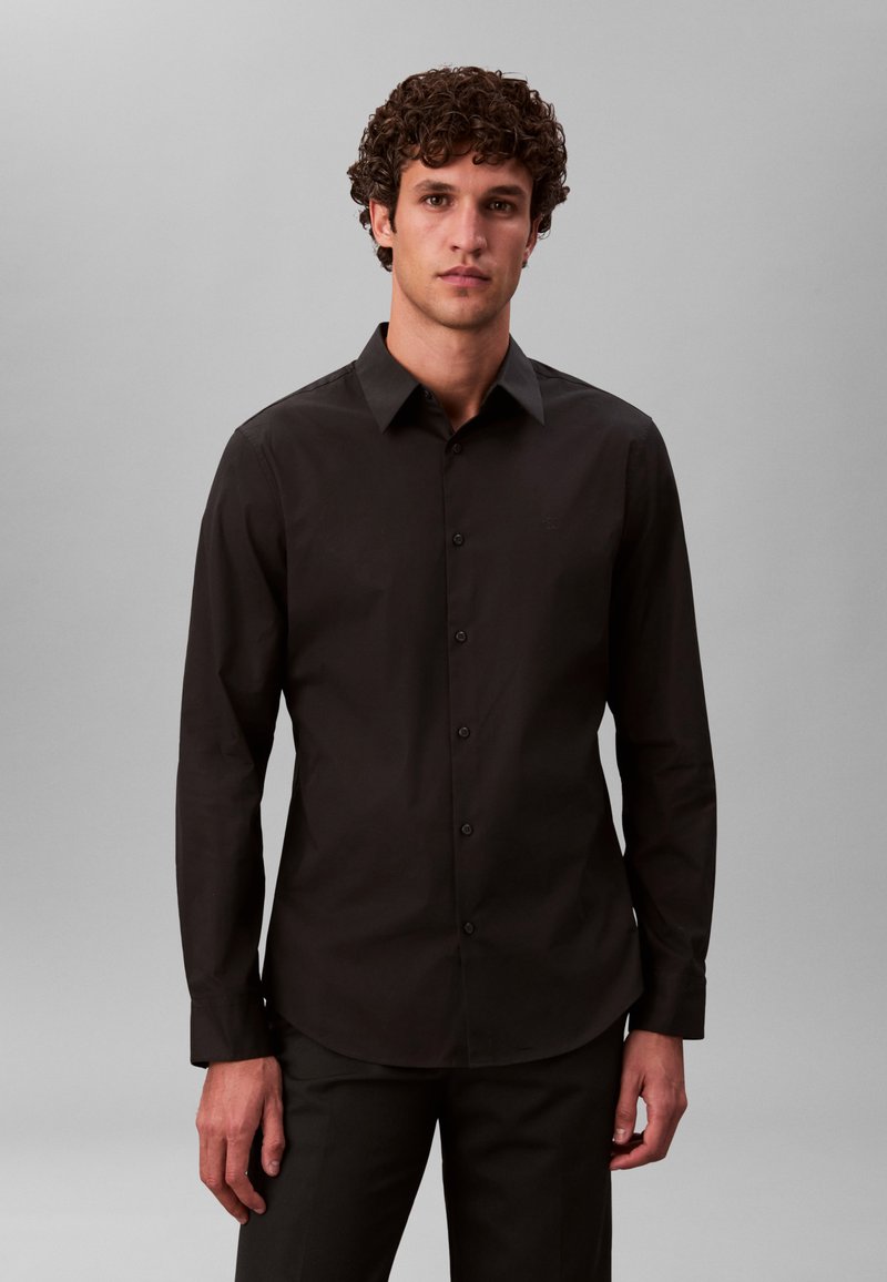 Čierna košeľa na gombíky z hladkej tkaniny, s ostrým golierom a dlhými rukávmi v úzkom strihu. Jemný detail loga na hrudi.