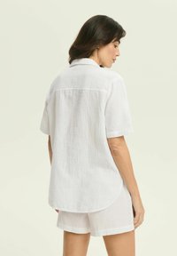 White short-sleeve button-up shirt made of lightweight linen. Features a curved hem and subtle texture, styled with matching shorts.