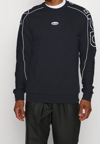 Sudadera negra con ribetes blancos en los hombros, cuello redondo y un pequeño logo en el pecho. Corte casual con mangas largas.