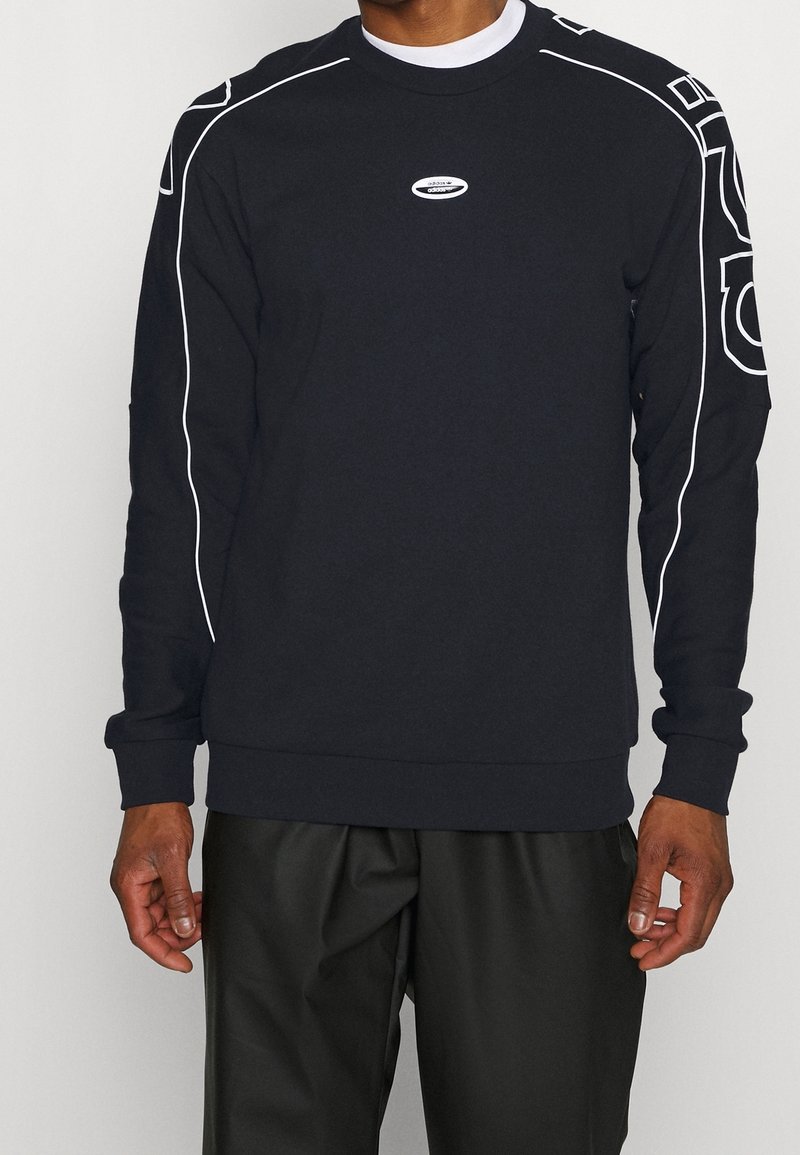Sudadera negra con ribetes blancos en los hombros, cuello redondo y un pequeño logo en el pecho. Corte casual con mangas largas.