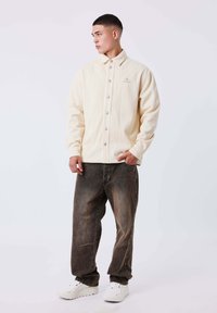 Chaqueta de rayas color beige claro con botones plateados, combinada con jeans oscuros desgastados con botones laterales y zapatillas blancas.
