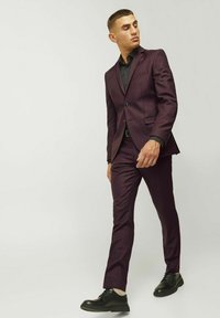 Costume bordeaux avec un design ajusté, veste à un bouton et pantalon droit, associé à des chaussures noires et une chemise noire.