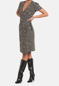 Žena ve květinových šatech černobílé barvy po kolena, s krátkými rukávy a černými vysokými kozačkami, s rukama na bocích.