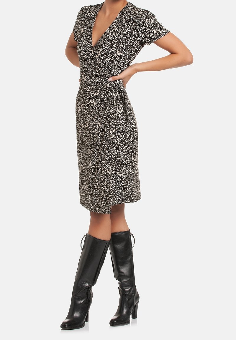 Žena ve květinových šatech černobílé barvy po kolena, s krátkými rukávy a černými vysokými kozačkami, s rukama na bocích.