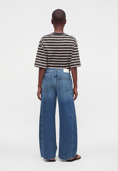 Personne de dos, portant un t-shirt rayé noir et beige, un jean bleu à jambes larges et des sandales noires sur un fond uni.