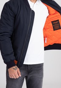 Muž v námořnickém bomberu, který odhaluje jasně oranžovou vypodšívku s prošíváním, černou nášivkou, bílém polo tričku, tmavých džínách a tetováním na ruce.