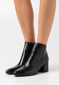 Botas de tobillo de cuero negro con punta afilada, tacón cuadrado y acentos de tachuelas a lo largo del costado. Textura suave con un diseño elegante.