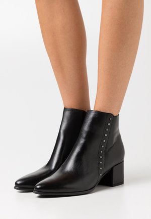 Botas de tobillo de cuero negro con punta afilada, tacón cuadrado y acentos de tachuelas a lo largo del costado. Textura suave con un diseño elegante.