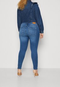 Žena v bodech modrých džín a tmavé denimové bundě, stojící zády k fotoaparátu na jednobarevném bílém pozadí.