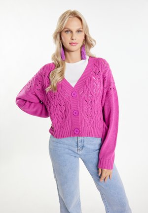 Heldere roze gebreide cardigan met een V-hals, openwerk patroon en drie grote knopen, gecombineerd met lichtblauwe jeans. Een soepele textuur en een ruime pasvorm.