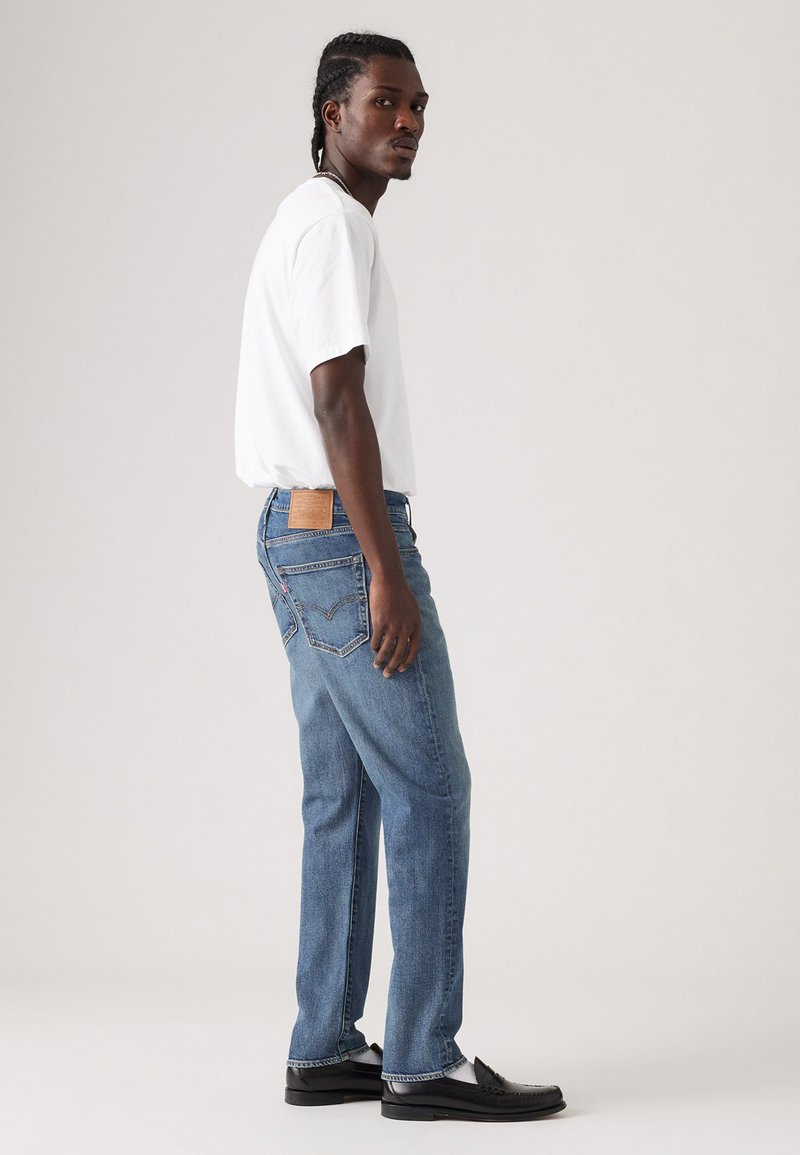 Jean en denim bleu clair avec une coupe droite, présentant une pièce en cuir marron au niveau de la taille et des détails de couture subtils. Associé à un T-shirt blanc.