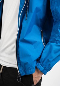 Blå letvægtsjakke lavet af glat, vandafvisende stof, med en lynlåslomme, elastiske manchetter og hætte med snor.