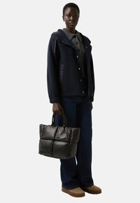 Vrouw in een marineblauwe jas, grijze knooptrui, donkere jeans, beige schoenen, met een zwarte gewatteerde schoudertas met een ronde logotag.