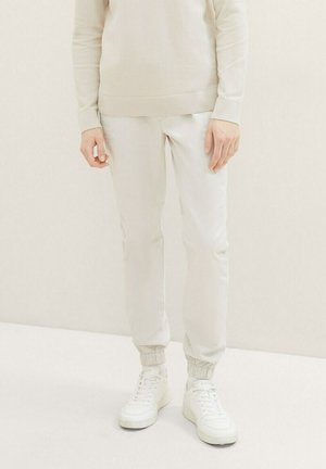 Joggers color beige claro con una cintura y puños elásticos, combinados con zapatillas blancas. El material parece suave con un ligero brillo.