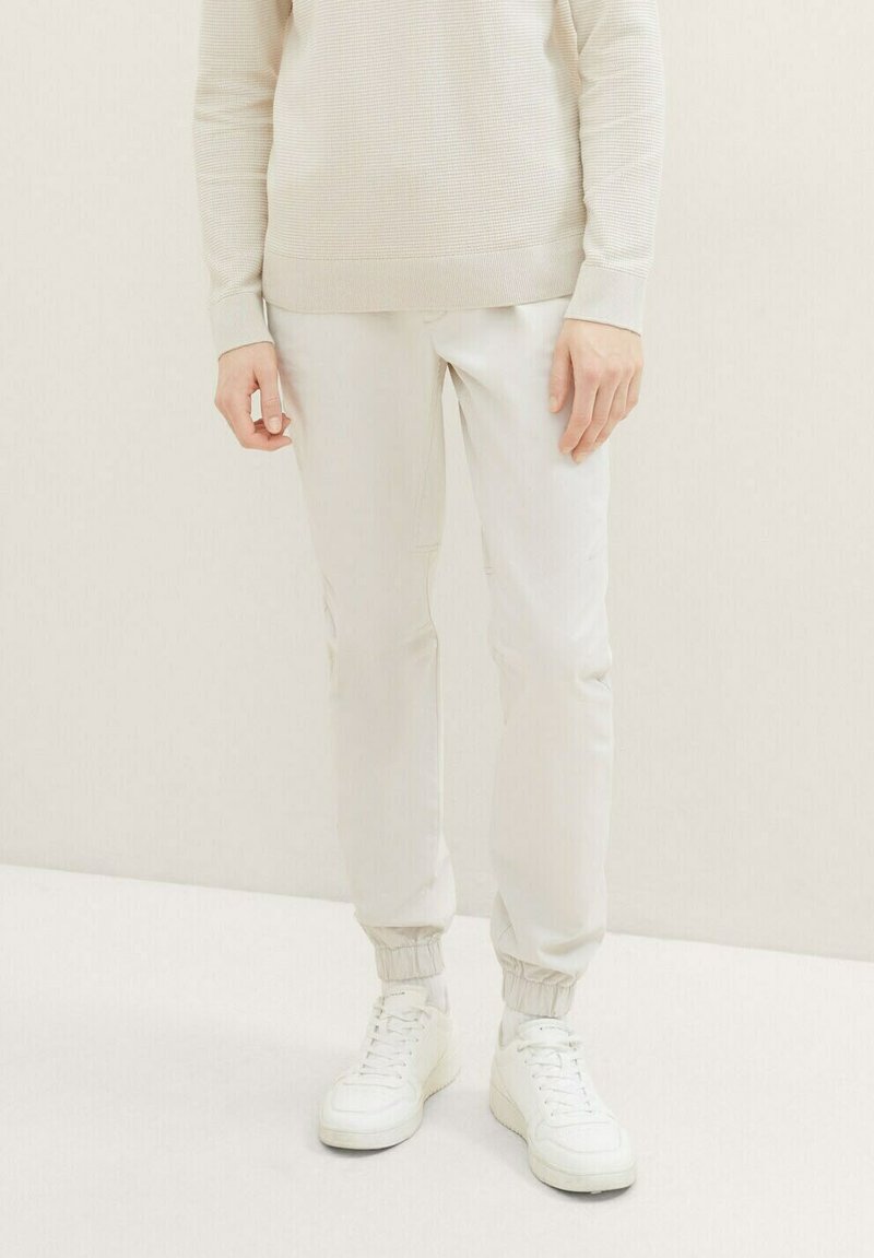 Joggers color beige claro con una cintura y puños elásticos, combinados con zapatillas blancas. El material parece suave con un ligero brillo.