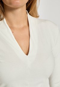 Hvid rippet top med dyb V-udskæring, der har en subtil tekstur og et tætsiddende design, der følger kroppens former.