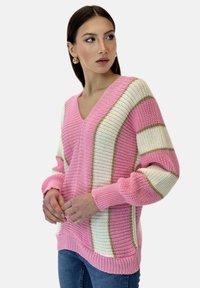 Pull en V rose avec des motifs rayés blancs et dorés, texture tricotée et manches longues, porté sur un jean bleu.