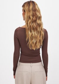 Ruskea pitkähihainen istuva paita, jossa on neliön muotoinen kaula-aukko ja rypytysdetailia, yhdistettynä vaaleisiin räätälöityihin housuihin.