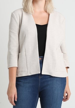 Mujer con chaqueta beige claro abierta al frente y mangas tres cuartos, camiseta negra y vaqueros azules sobre un fondo liso.