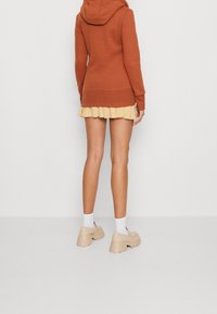 Felpa con cappuccio marrone dal taglio rilassato, abbinata a una gonna gialla con balze. Moda che indossa scarpe con piattaforma color crema e calze bianche.