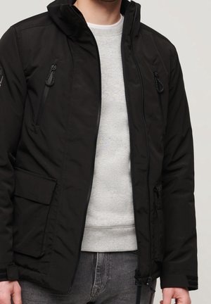 Homme portant une veste noire zippée par-dessus un pull gris clair et un jean gris foncé devant un fond clair uni.
