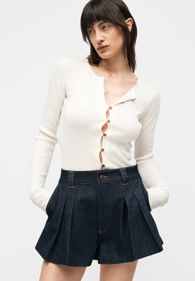 Γυναίκα με λευκή ριμπ πλεκτή μπλούζα με μακριά μανίκια και καφέ κουμπιά και σκούρα πλισέ σορτς με ψηλή μέση, χέρια στις τσέπες, κοιτάζει αριστερά.