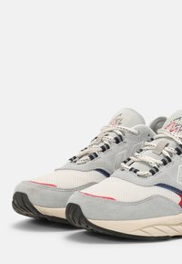 Grå och vita sportskor i mesh och mocka, med blå och röda accenter, texturerade sulor och kontrasterande snören.