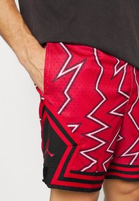 Röda idrottsshorts med svarta accenter, med ett zigzag-mönster och sidofickor. Tillverkade av andningsbart nätmaterial.