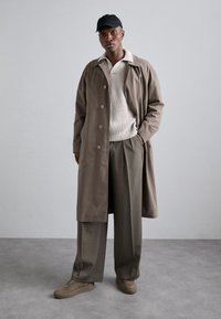 Cappotto beige lungo con bottoni, maglione di lana color crema, pantaloni olive larghi e sneakers beige. Cappellino nero completa la palette di colori neutri.